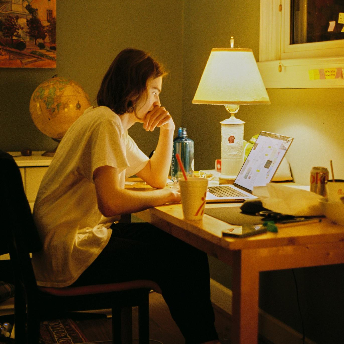 A person working late at a cluttered desk, lit by a warm lamp while looking at a laptop screen.