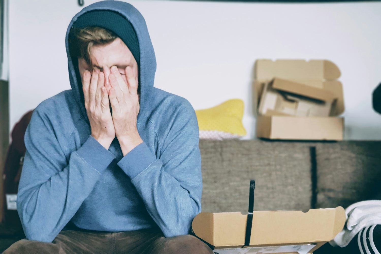 A person in a blue hoodie sitting on a couch with their face in their hands, appearing stressed or overwhelmed, with open cardboard boxes in the background.