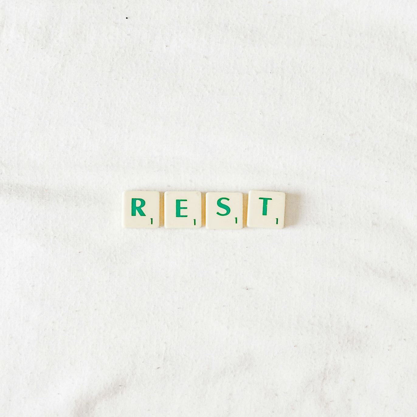 Scrabble-style letter tiles spelling the word “REST” on a soft white fabric background.