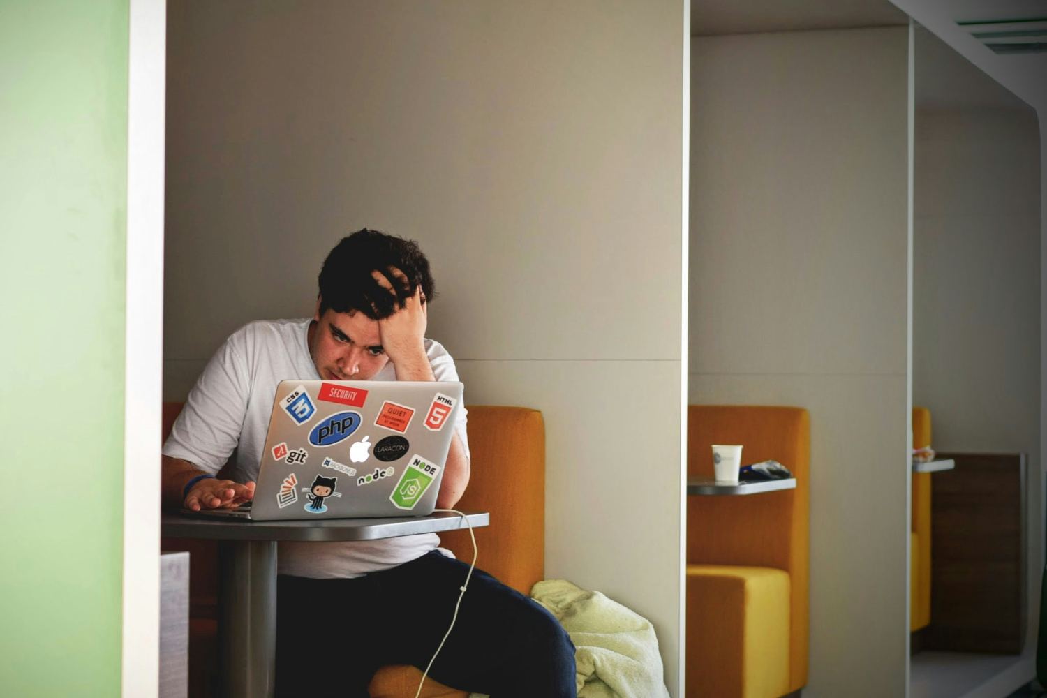 A person sitting in a small booth, looking stressed while working on a laptop covered in programming stickers, with their hand on their head.