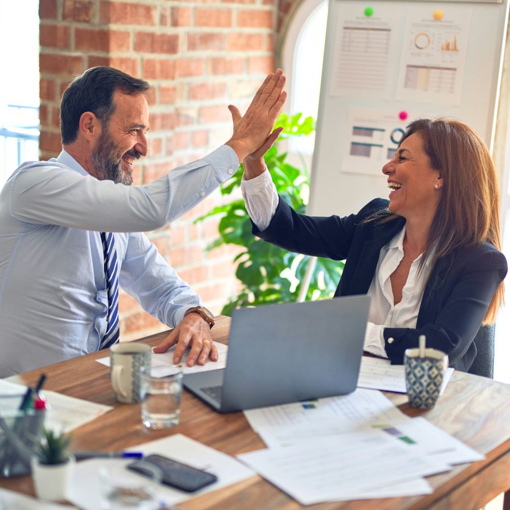 Two colleagues high-fiving at a desk with a laptop, celebrating success in an office.