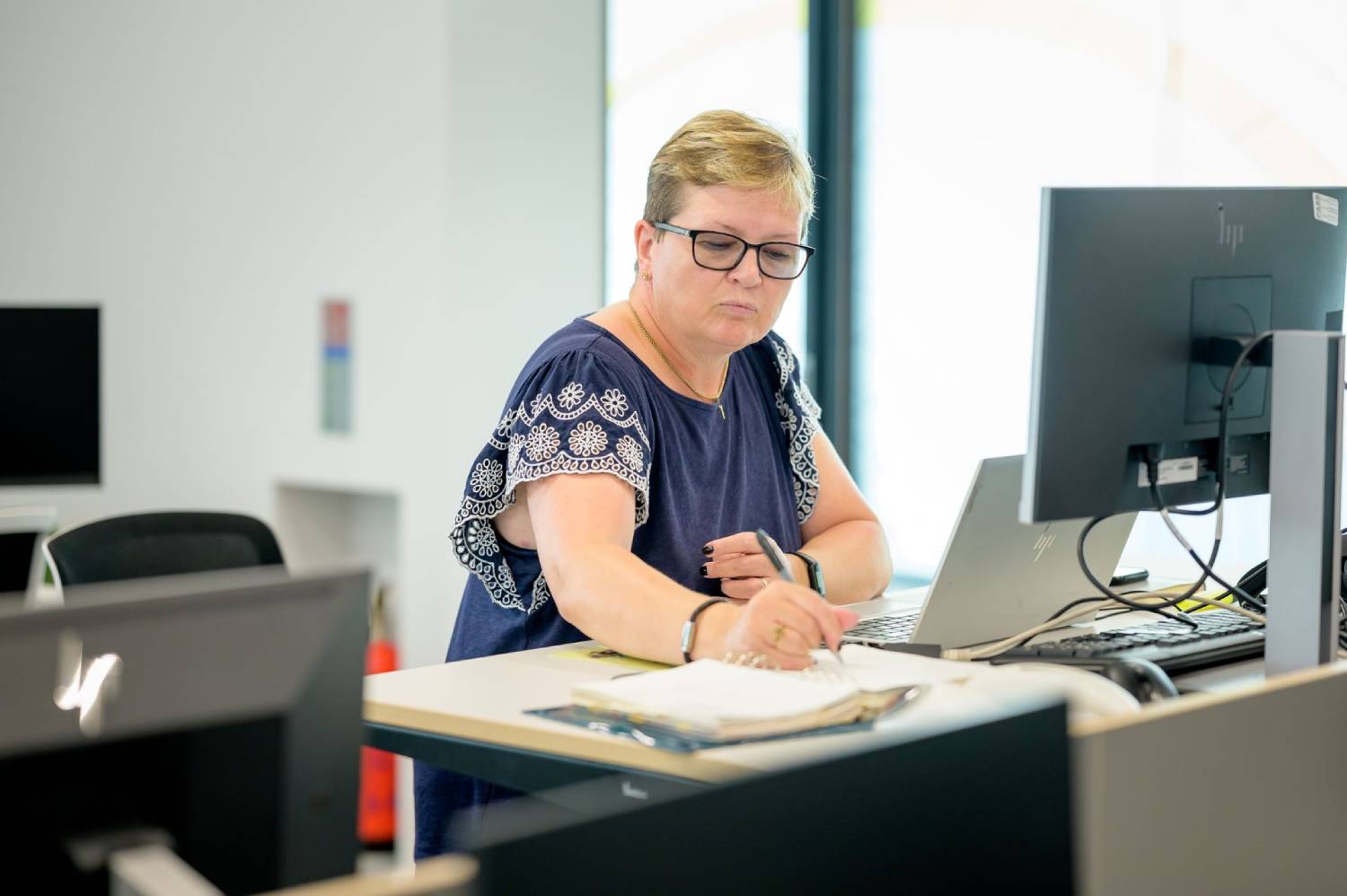 Woman working at a computer and writing notes at a desk in a bright office.