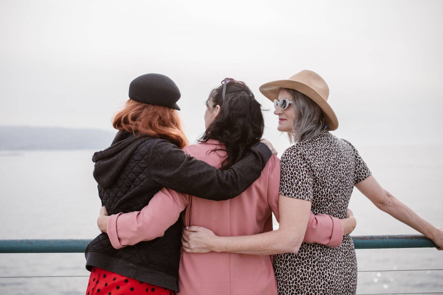 Three women standing arm in arm by a waterfront, wearing colourful outfits and hats while looking out over the water.