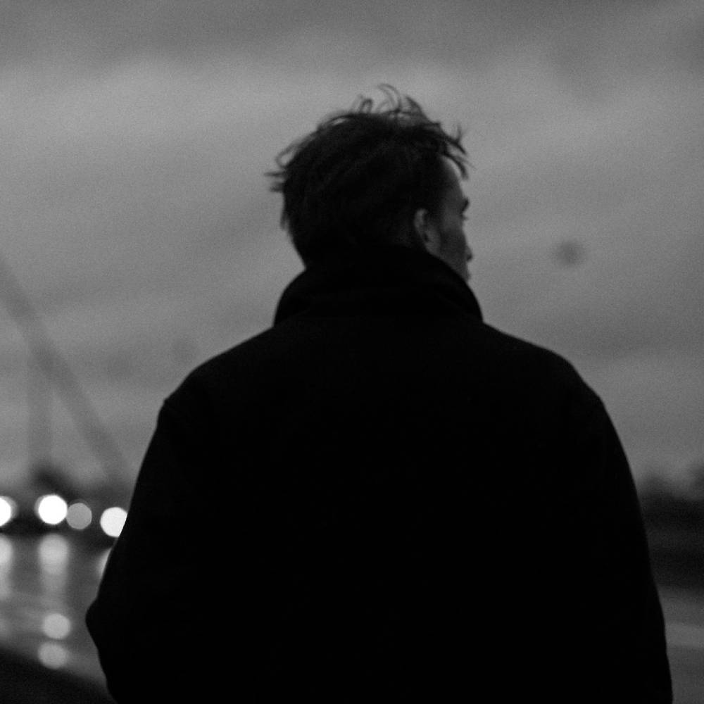 Black-and-white photo of a person in a dark coat walking along a waterfront at night, with blurred lights reflecting on the water.