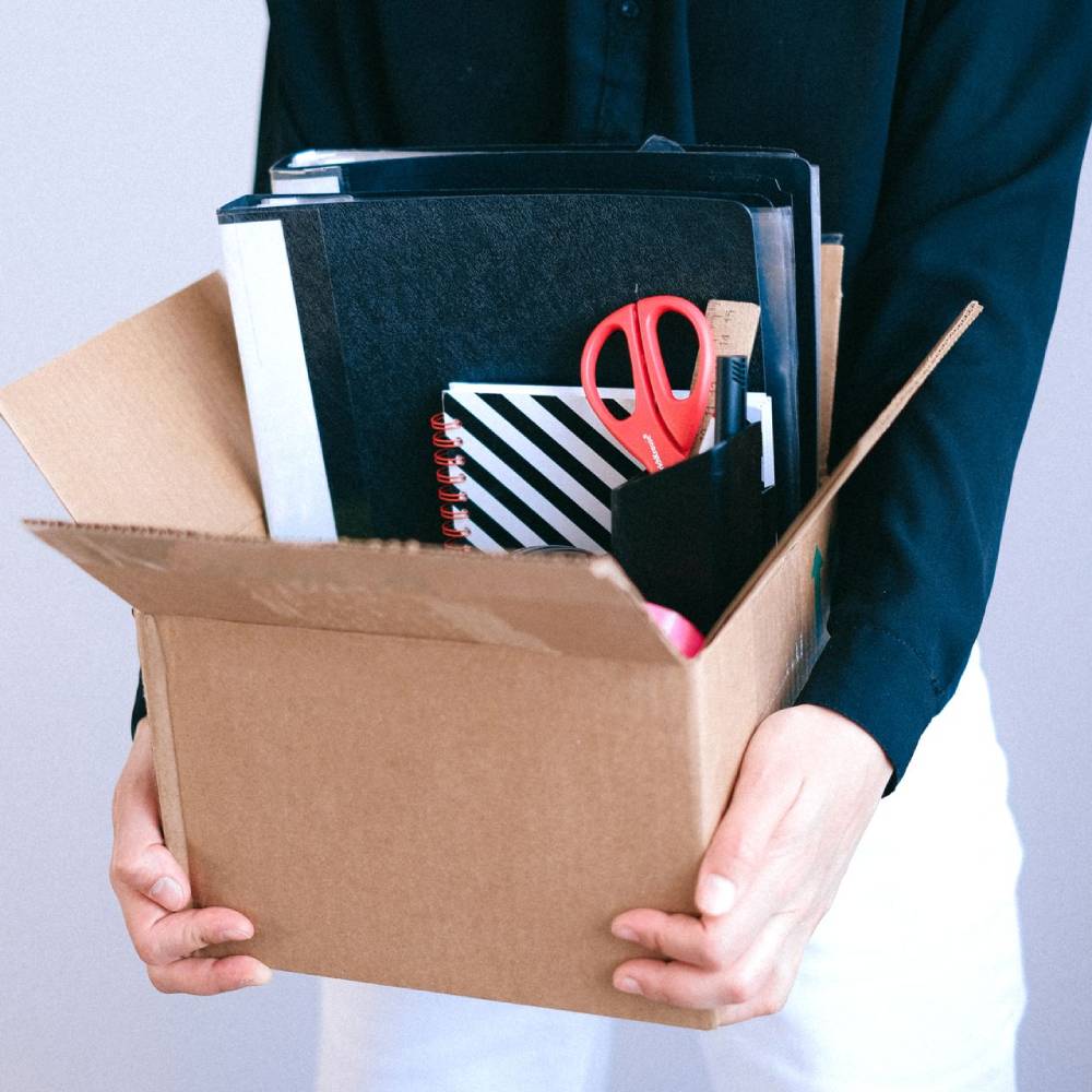 Person holding a box of office supplies, folders, notebooks, and scissors.