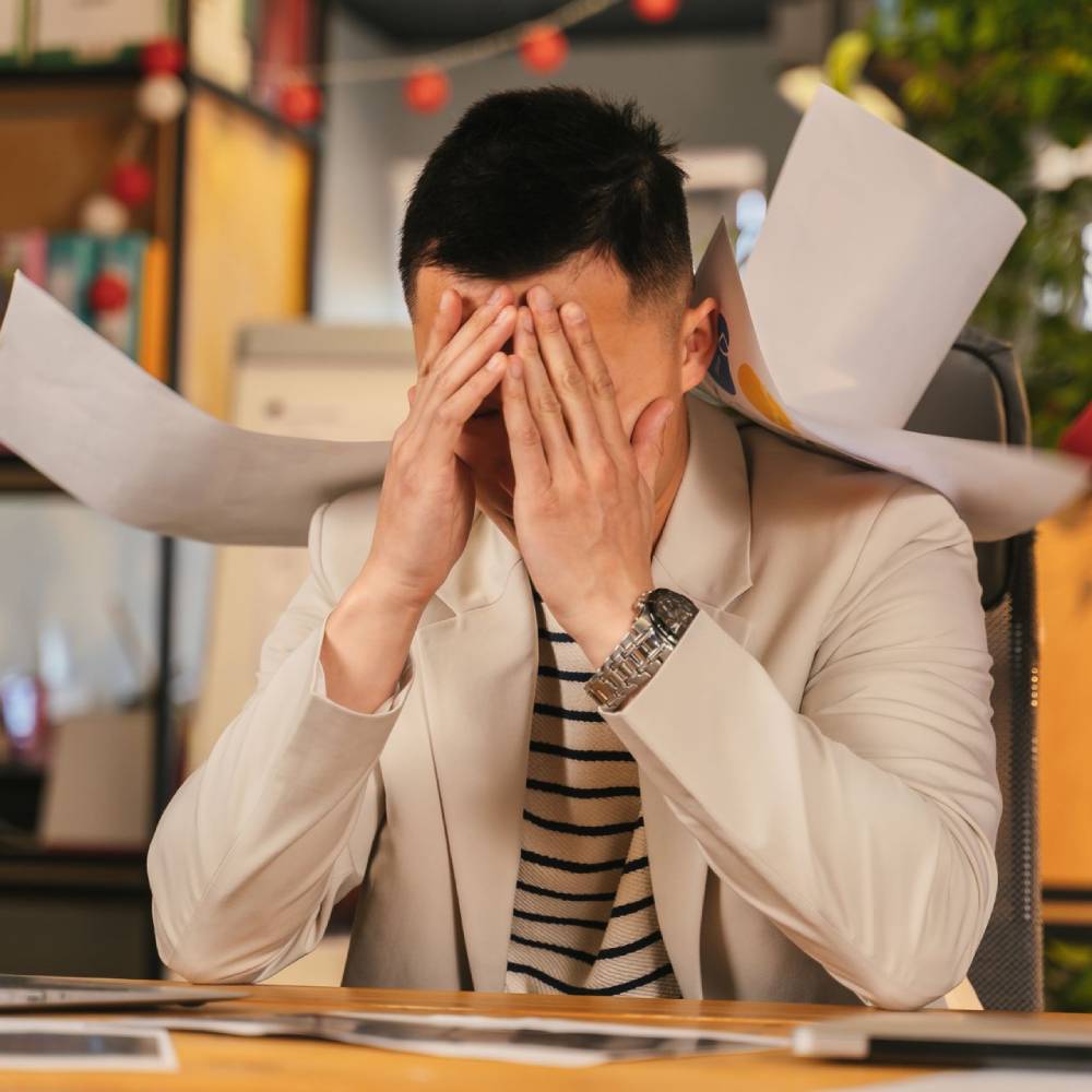 Stressed professional covering his face at a desk while papers are handed to him in a busy office.