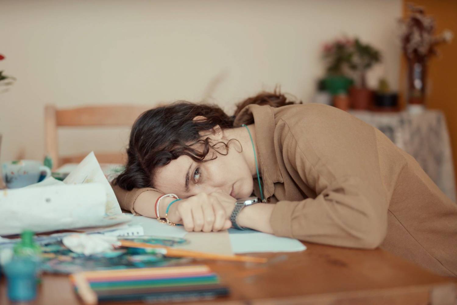 Tired woman resting her head on a desk surrounded by art supplies and papers.