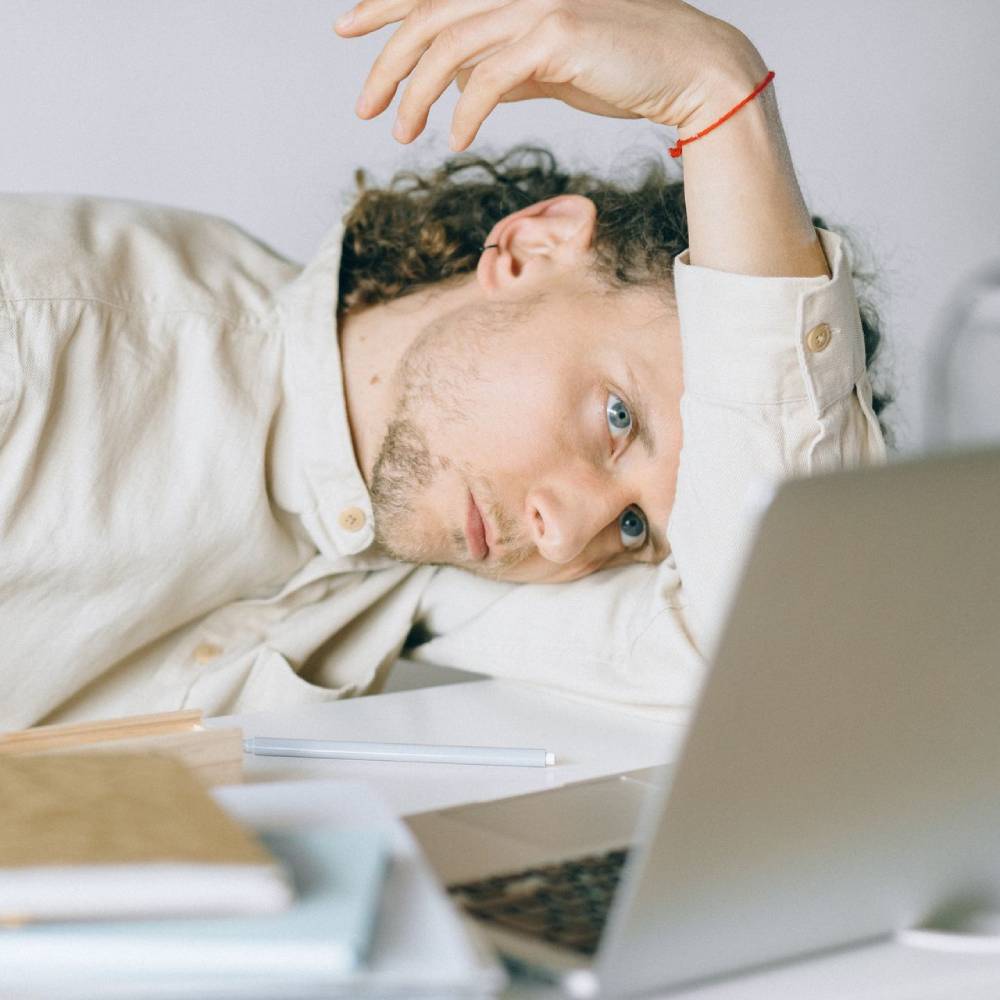 Tired man resting his head on a desk beside a laptop while working.