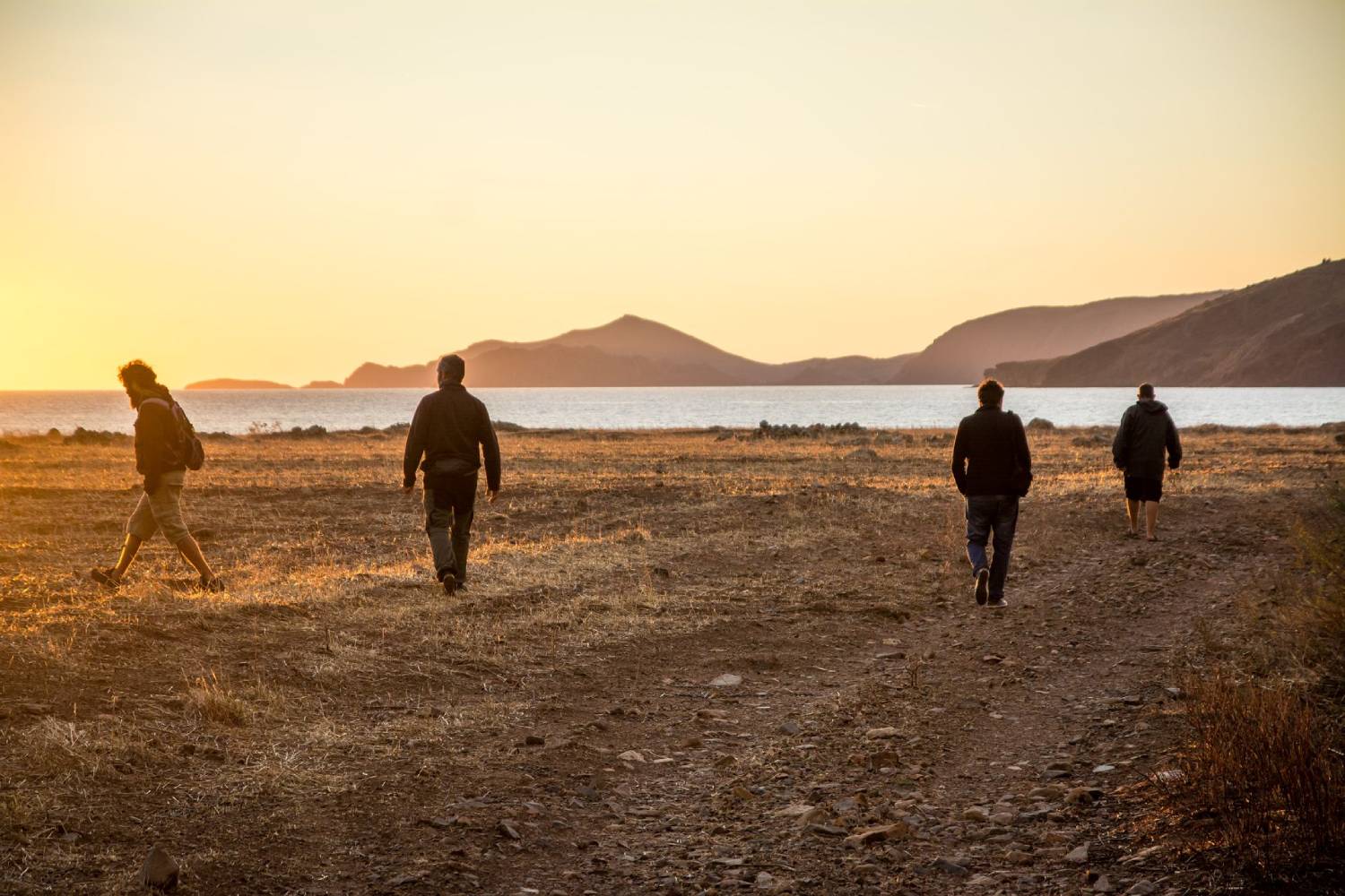 Four people walking across a coastal landscape toward the ocean at sunset, with mountains in the distance.