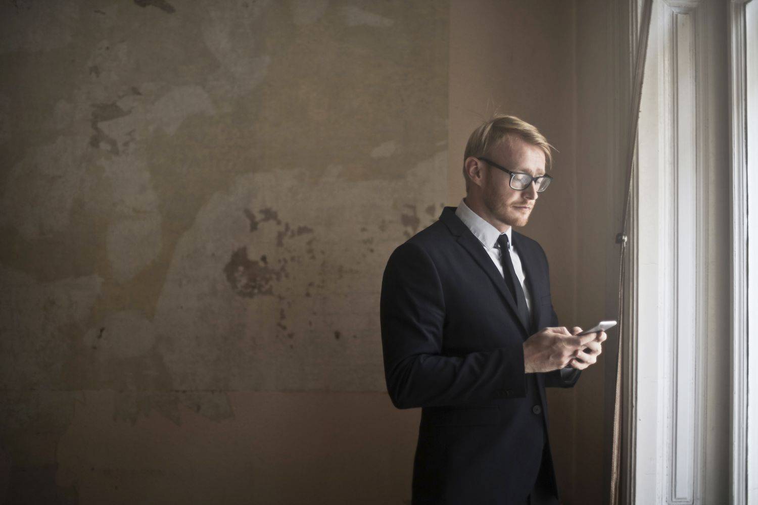 Businessman in a suit and glasses using a smartphone beside a large window.